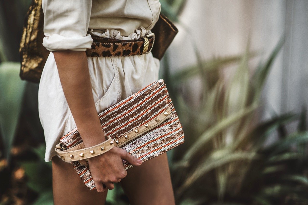 Person wearing beige romper with a leopard print belt, holding a woven clutch purse with studded accents, standing outdoors near plants.