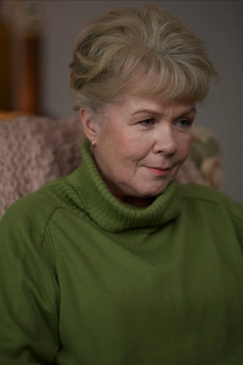 A middle-aged woman with short blonde hair wearing a green turtleneck sweater, sitting on a patterned couch in an indoor setting actor Sarah Woods carol hobday