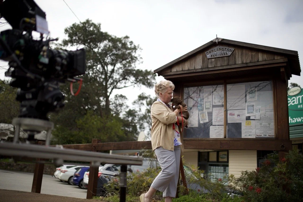 A woman holding a puppy in front of a town bulletin board in Darlington, established 1883, with cars parked in the background. actor Sarah Woods carol hobby