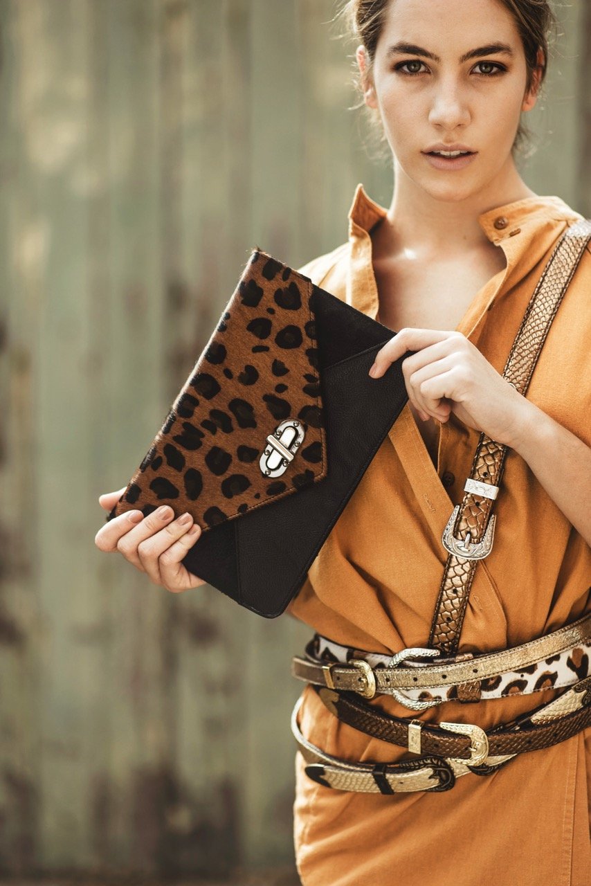 Woman outdoors holding a leopard print clutch purse, wearing a brown belt, and a sleeveless orange dress. She has a snake print handbag strap over her shoulder.