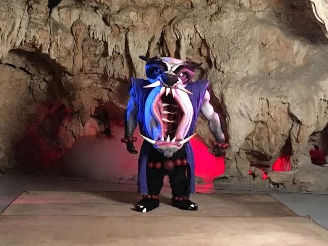 Person in a detailed monster costume with a large mouth and sharp teeth, standing inside a cave with rocks and red lighting.