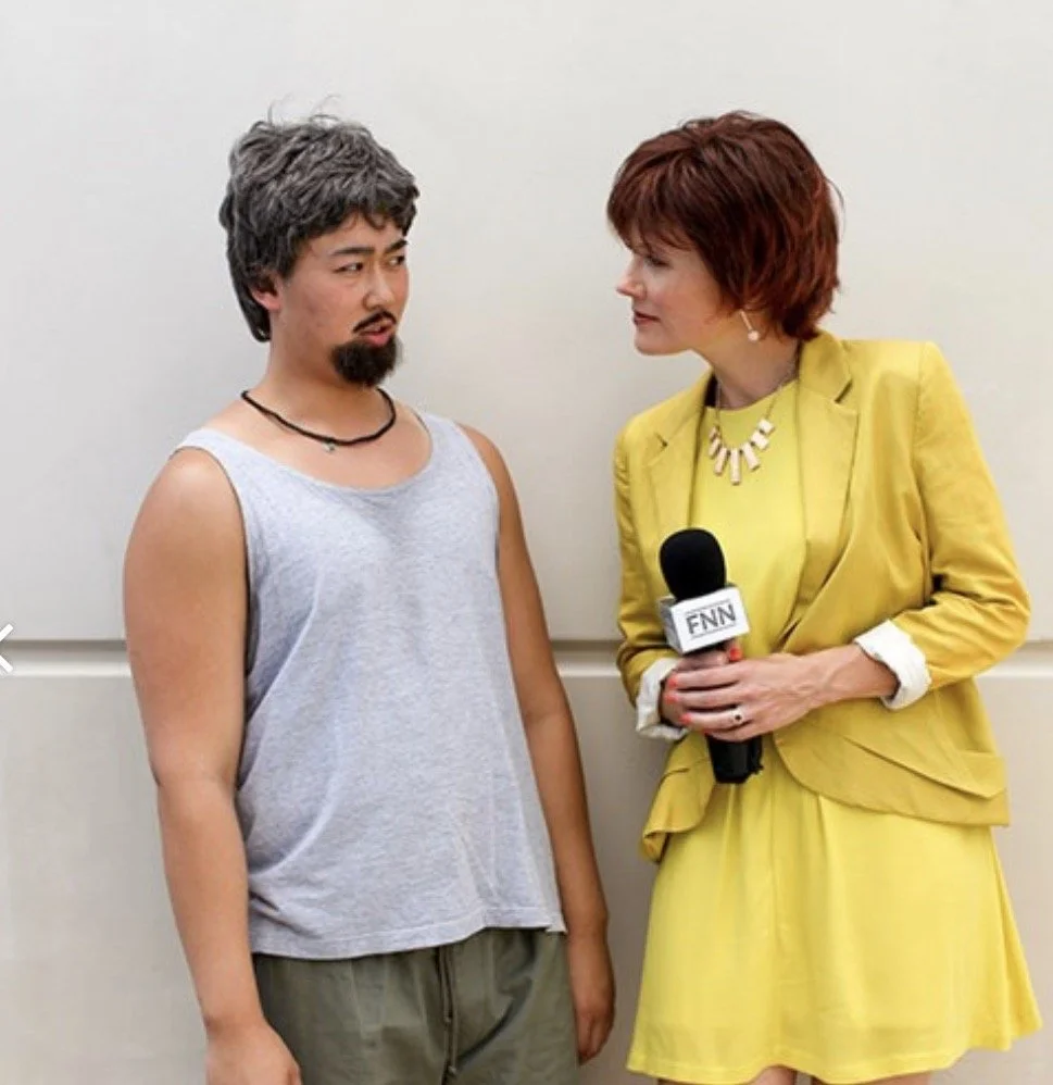 A woman in a yellow blazer holding a microphone interviewing a man dressed in a sleeveless shirt and casual pants, both standing against a plain wall.
