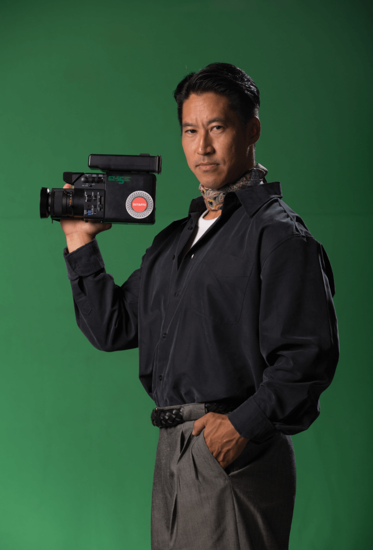 Man holding a professional video camera on his shoulder while standing against a green screen background.
