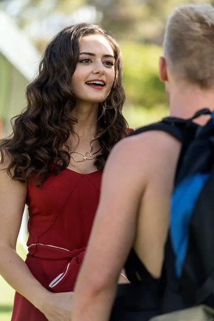 A young woman with long, curly brown hair wearing a red dress, talking to a young man with a backpack, outdoors on a sunny day.