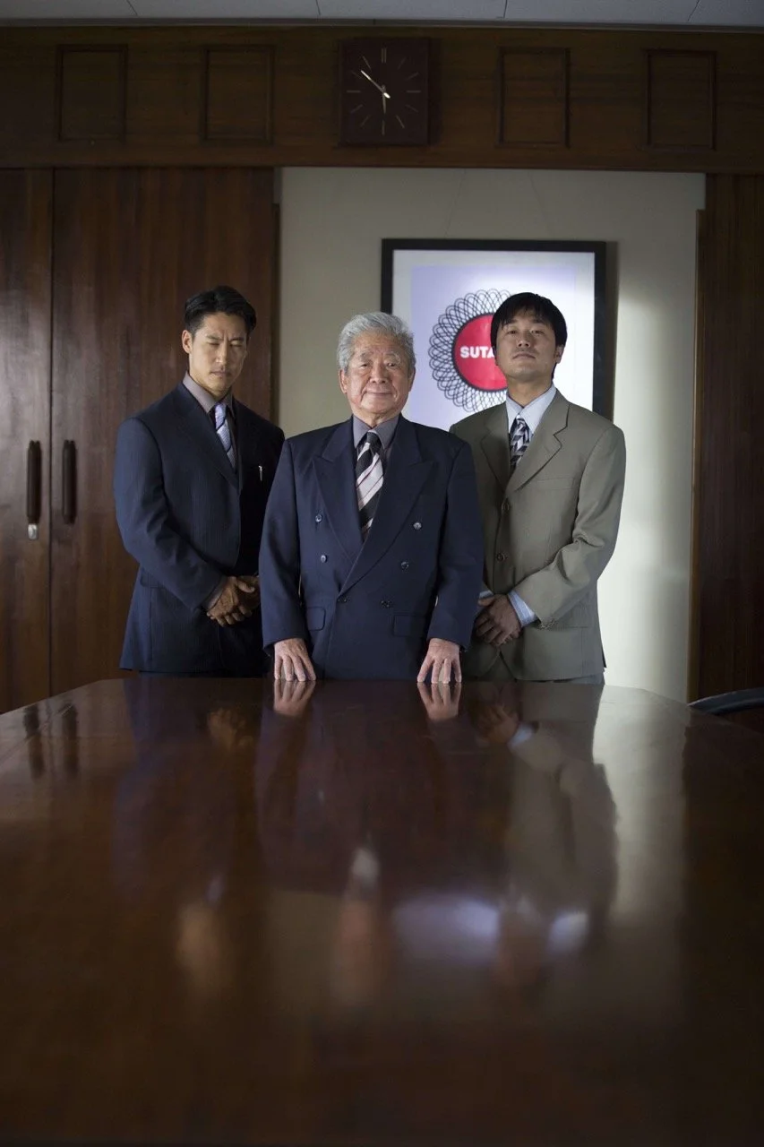 Three men in suits standing behind a polished conference table in a wood-paneled room with a clock and framed poster in the background  top knot detective  top knot detective