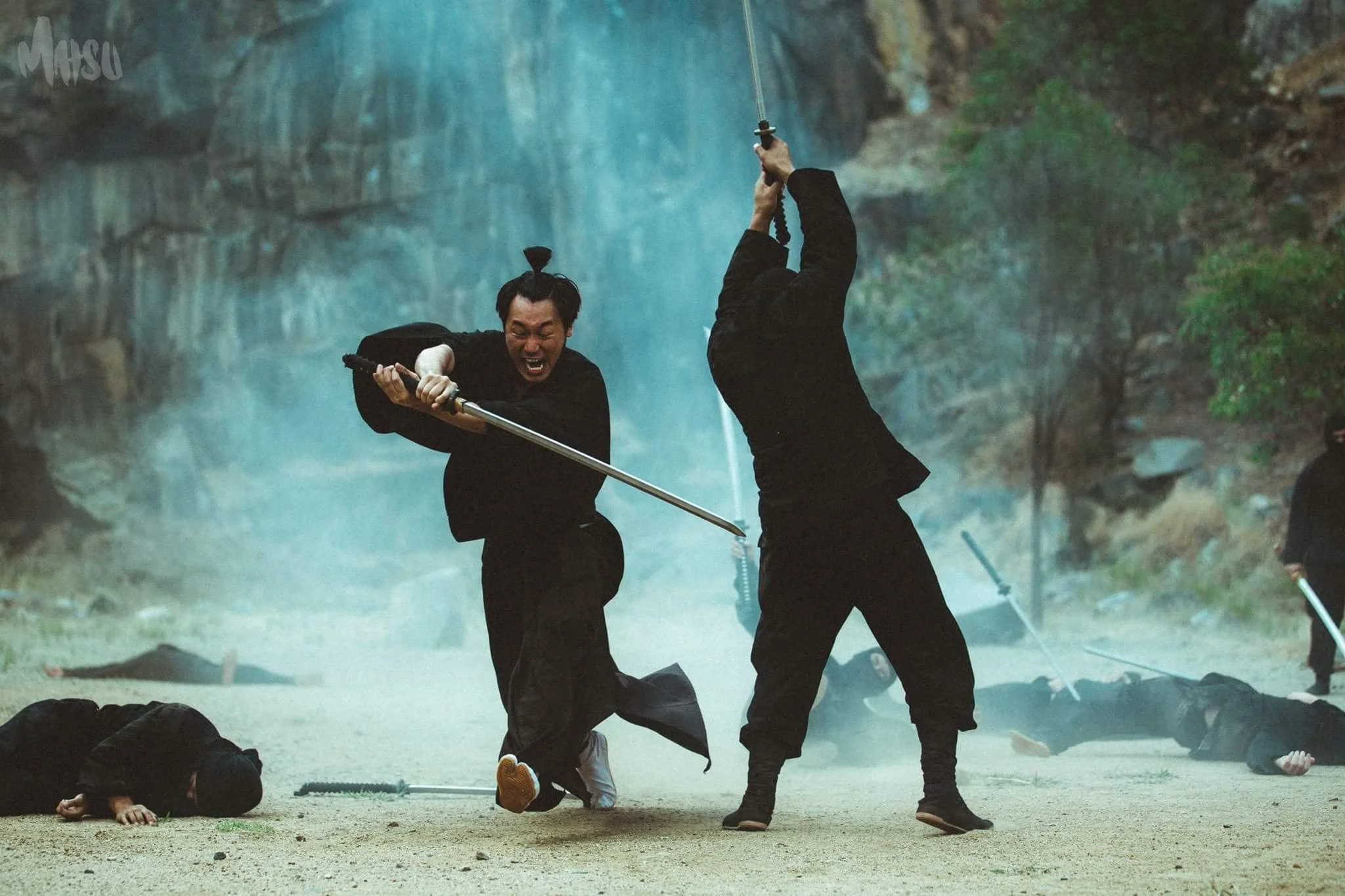 Two men with swords fighting in a historical battle scene, surrounded by others lying on the ground, set outdoors near a rocky cliff Top Knot Detective