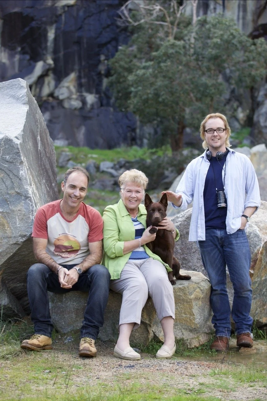 A group photo of three adults and a dog outdoors among rocks and trees. The woman in the center is holding a dark brown dog, and the man on the right is wearing glasses and has a camera hanging from his neck. The man on the left is sitting on a rock,