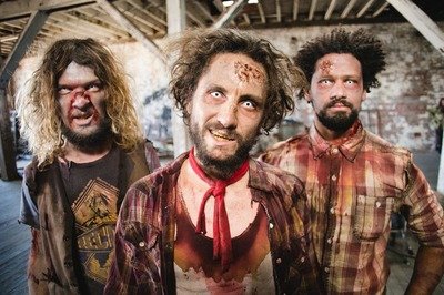 Three men with disheveled hair and zombie makeup in an industrial setting.