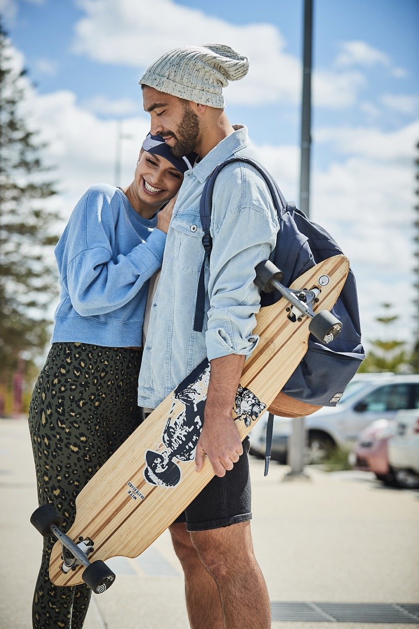 A smiling young woman hugging a bearded man holding a longboard skateboard outdoors on a bright, partly cloudy day, with parked cars and trees in the background.
