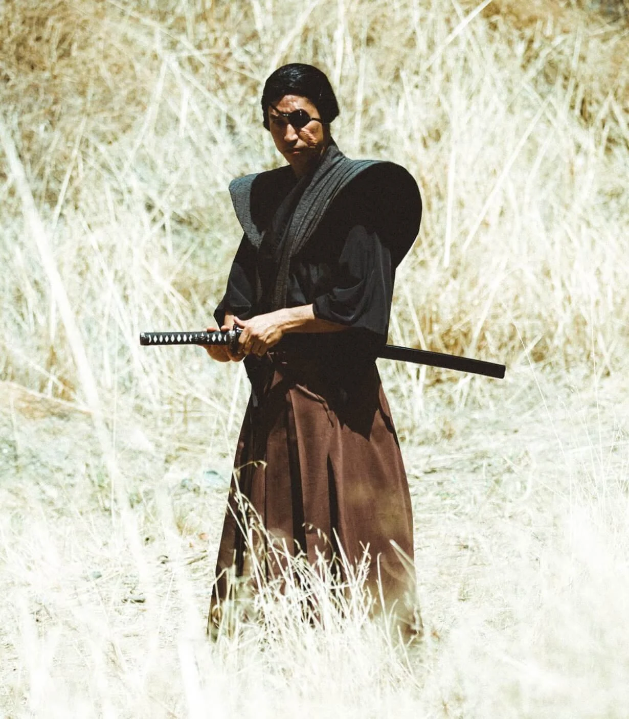 A person dressed as a samurai, holding a sword, standing in a field of tall grass or reeds.