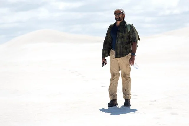 Man standing outdoors on a white sandy landscape, wearing sunglasses, a dark shirt, a green plaid jacket, beige cargo pants, and hiking boots, holding a water bottle in one hand and a phone in the other, with a backpack on his shoulders.