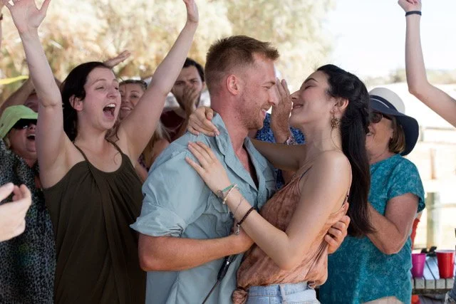 A group of people celebrating outdoors, with a man and woman embracing and others cheering around them.