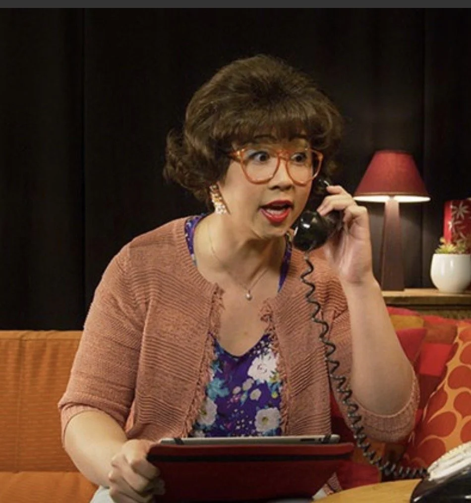 A woman with short curly hair and glasses, wearing a floral top and a cardigan, talking on a corded phone with a surprised or worried expression, sitting on a couch in a living room with a lamp and decorative pillow in the background.