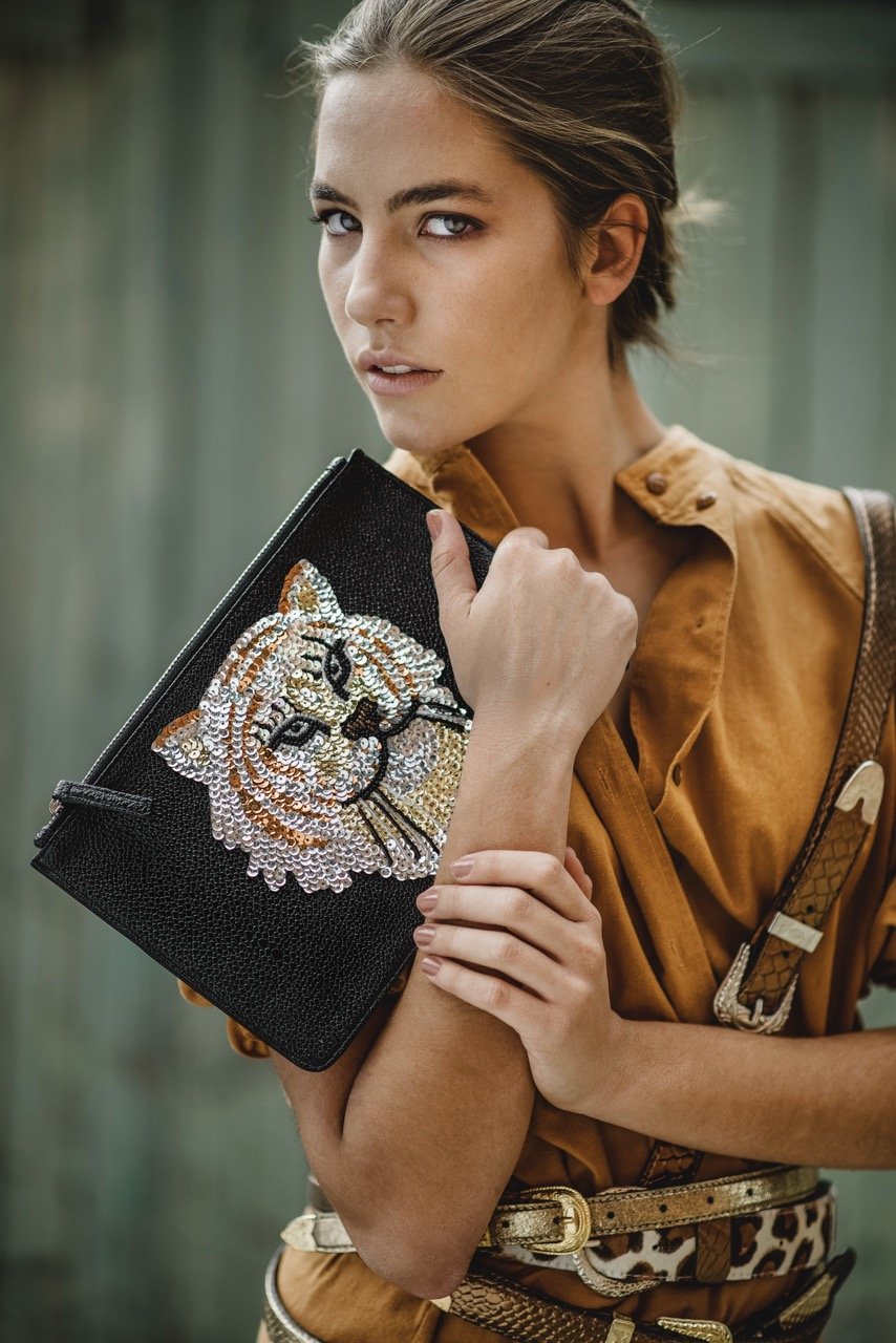 Young woman with short hair, wearing a tan button-up shirt, holding a black clutch purse with a sparkling tiger face design, standing outdoors with a green blurred background.