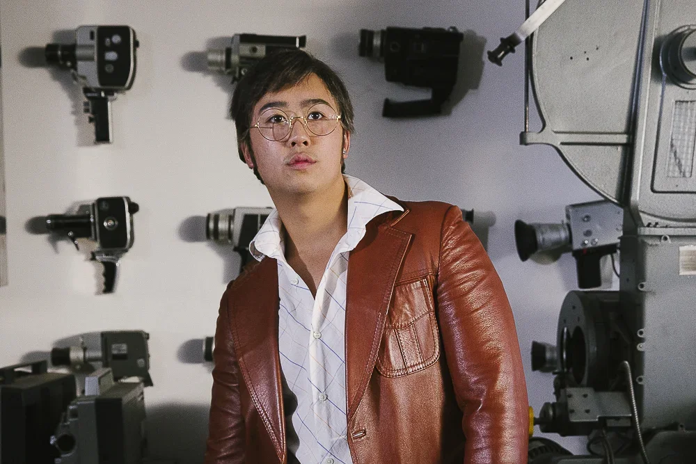 A man in glasses and a brown leather jacket stands in front of a wall with various gun-shaped objects and machinery.