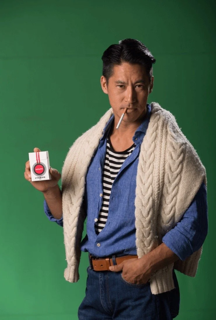 A man wearing a striped shirt and unbuttoned blue denim shirt, with a cream cable-knit sweater draped over his shoulders, holding a small matchbox, with a cigarette in his mouth, standing against a green background.