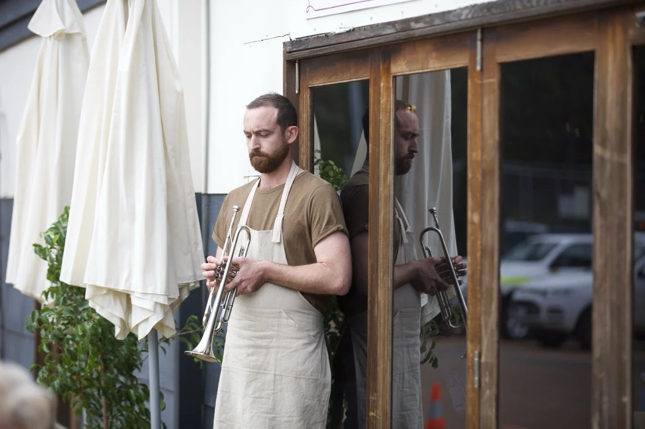 A man with a beard wearing a brown T-shirt and beige apron standing outside a building, holding a trumpet, and looking down pensively, with his reflection visible in a glass door beside him.
