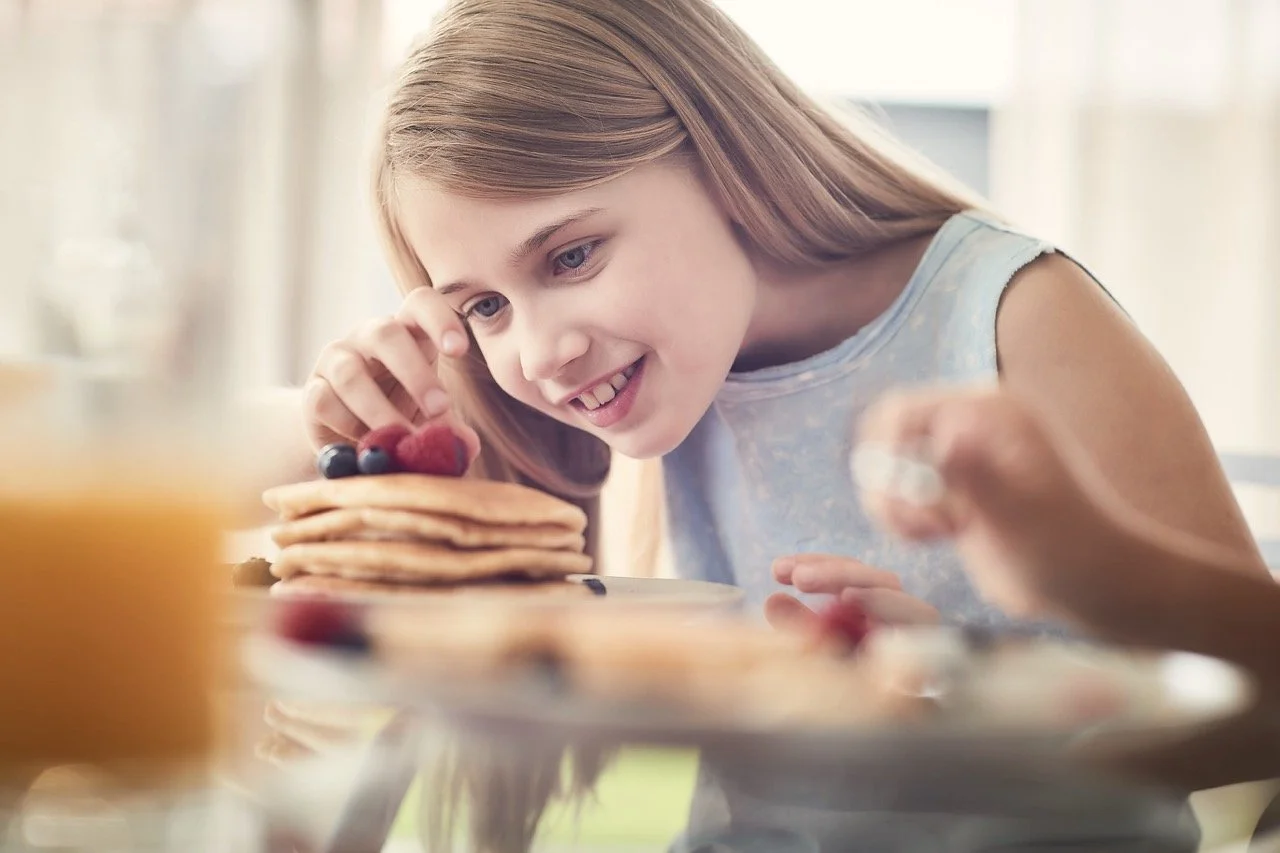 A young girl with blonde hair smiling and reaching for a stack of pancakes topped with berries at breakfast.