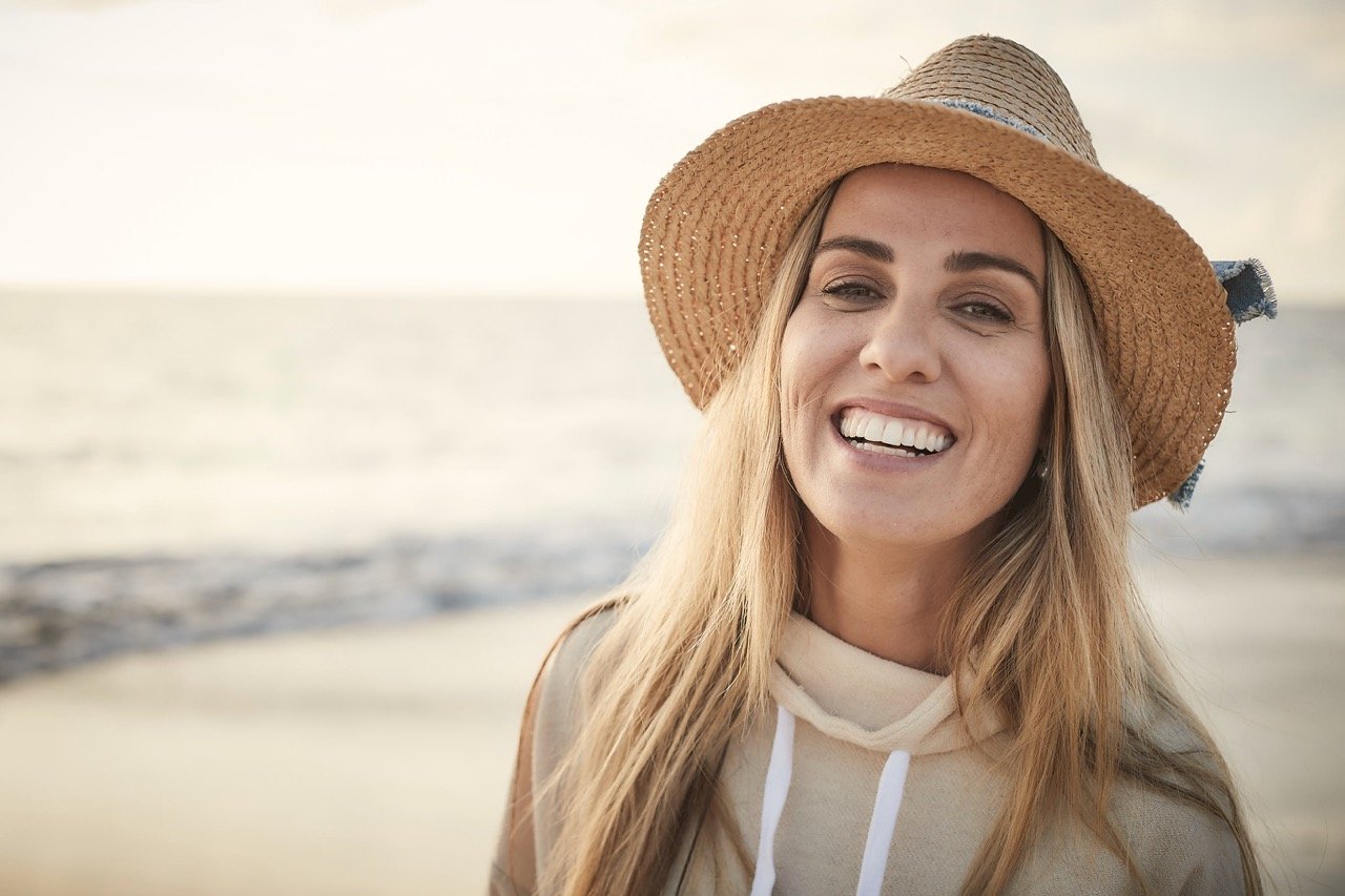 A woman smiling at the camera at the beach, wearing a wide-brimmed straw hat and a beige hoodie.