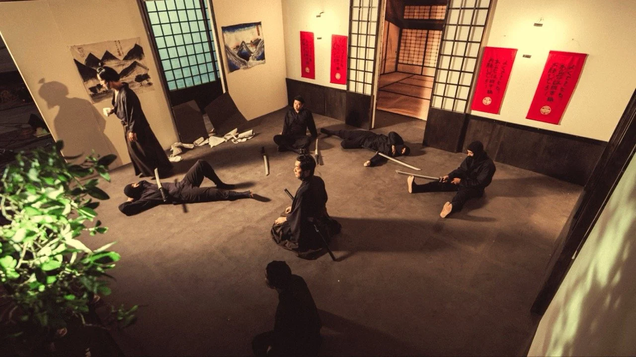 People practicing a martial arts demonstration inside a traditional Japanese room with sliding shoji doors and Japanese artwork on the walls.