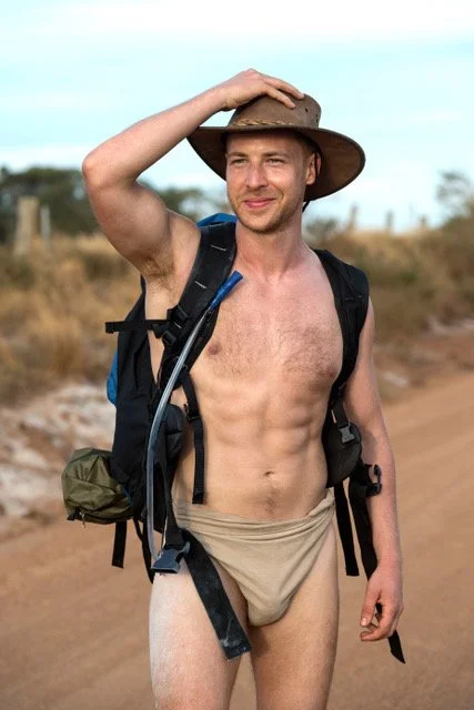 A shirtless man wearing a wide-brimmed hat and a backpack, standing outdoors with a dirt path and grassy landscape in the background.