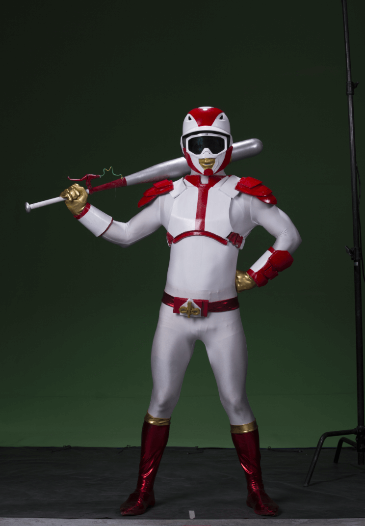 A person dressed in a superhero costume resembling a baseball player with a helmet, goggles, white and red suit, gold gloves, and red boots, holding a baseball bat over their shoulder in front of a green background.