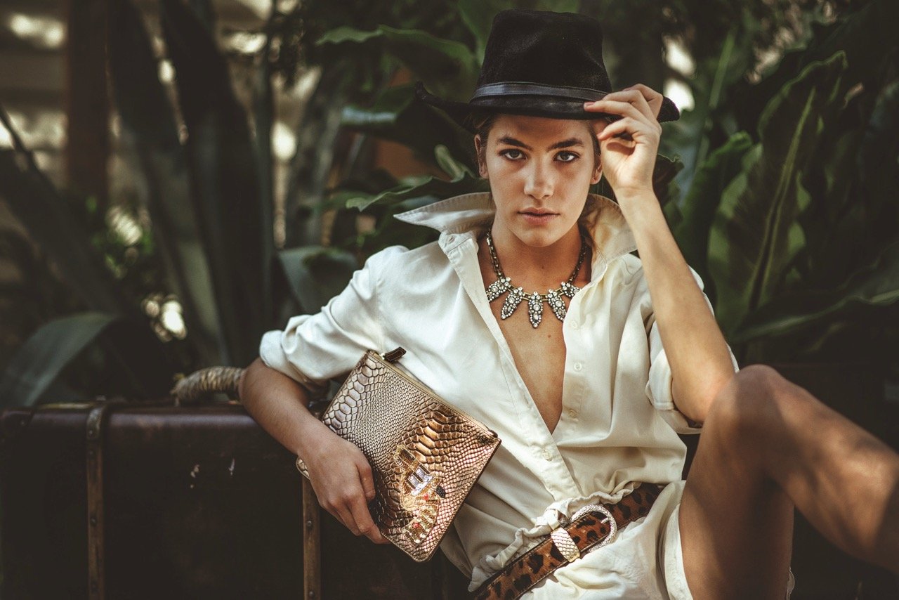 A woman sitting outdoors on a bench, wearing a white shirt, leopard print belt, and holding a gold snakeskin clutch purse. She is adjusting a black hat and looking directly at the camera, with large green plants in the background.