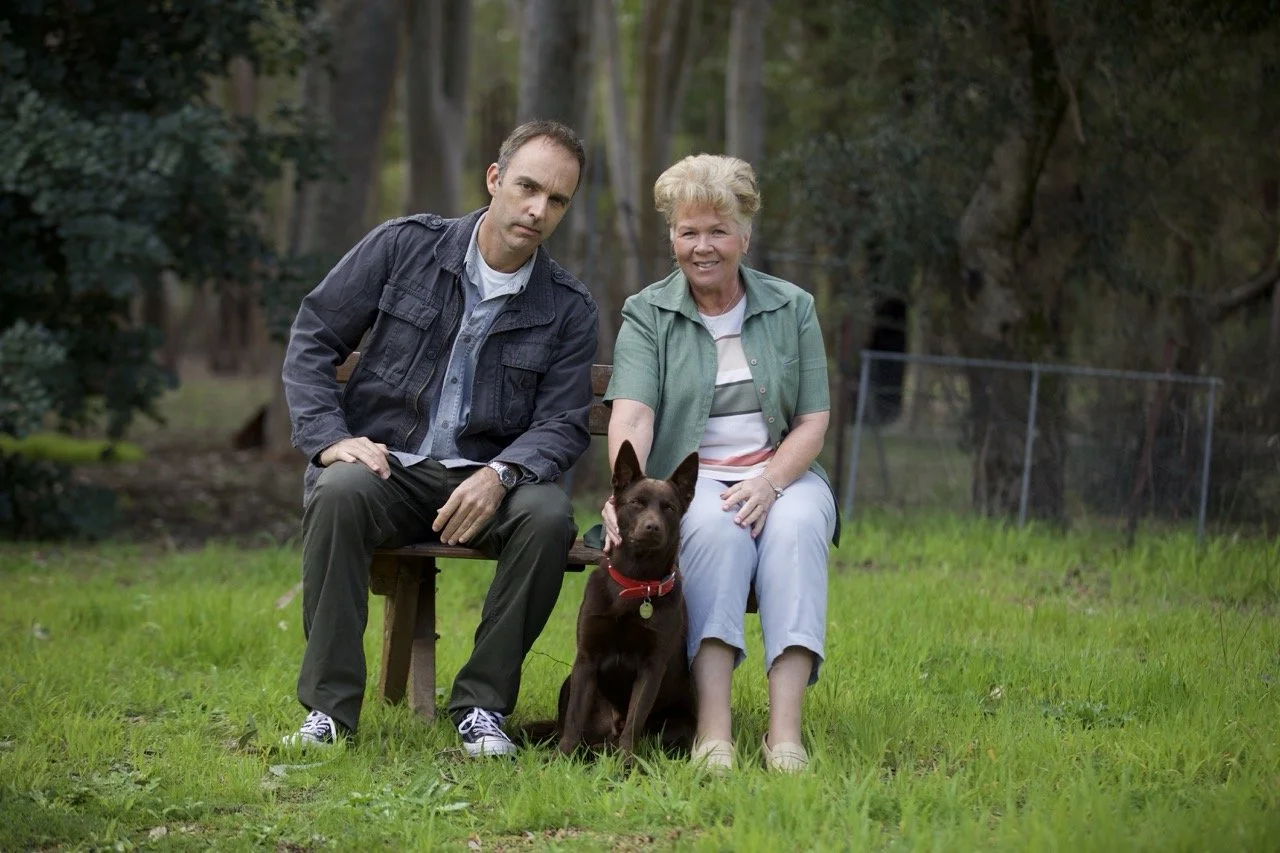 A man and a woman sit on a bench outdoors with a brown dog between them. The man wears a dark jacket and pants, and the woman wears a green shirt and light-colored pants. They are in a grassy area with trees in the background.
