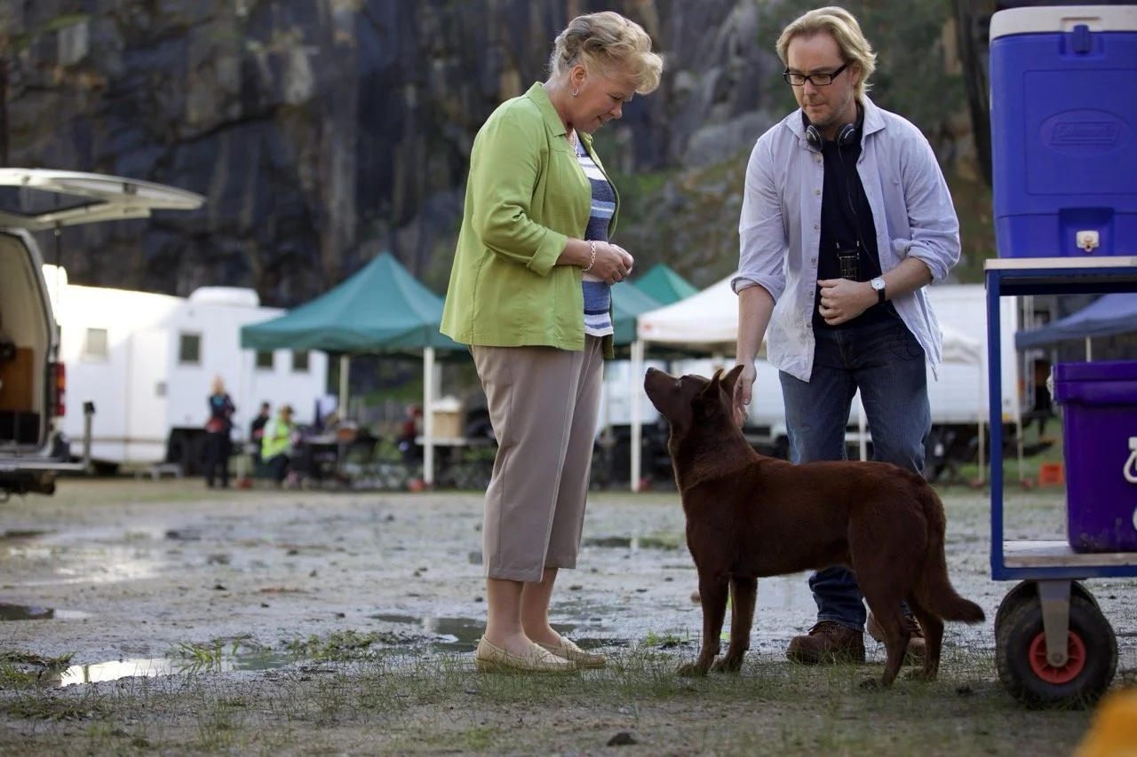 A woman and a man are standing outdoors with a brown dog. The woman is wearing a green jacket and beige pants, and the man has glasses and is wearing a light blue shirt. They appear to be at a campground with tents and RVs in the background.