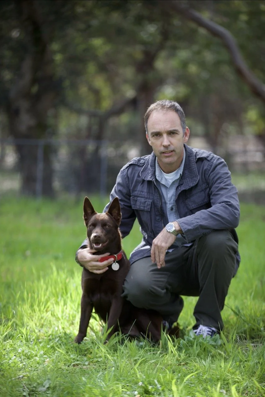 A man kneeling on grass outdoors next to a brown dog with pointed ears and a red collar, with trees in the background.