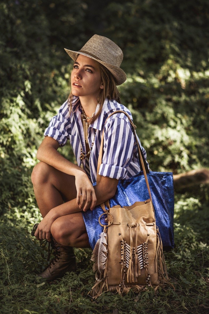 A young woman in a striped shirt, hat, and jewelry, squatting outdoors with backpacks, surrounded by greenery.