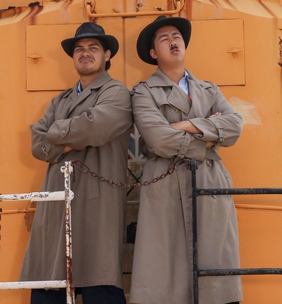 Two men in trench coats and black wide-brimmed hats standing back-to-back with arms crossed, posing in front of a yellow-orange painted metal surface.