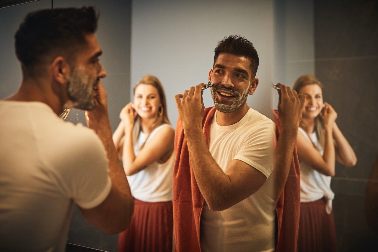 Two men in a bathroom are shaving, with women smiling and preparing behind them, reflected in the mirror.