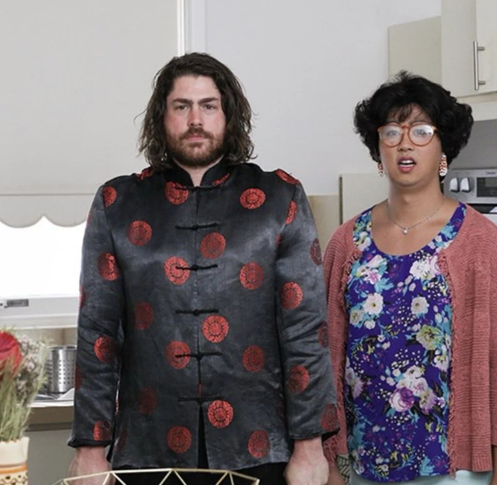 Two people standing in a kitchen, one with long, dark, wavy hair in a black and red traditional Chinese-style jacket, and the other with short, curly hair, glasses, and a floral shirt with a pink cardigan.