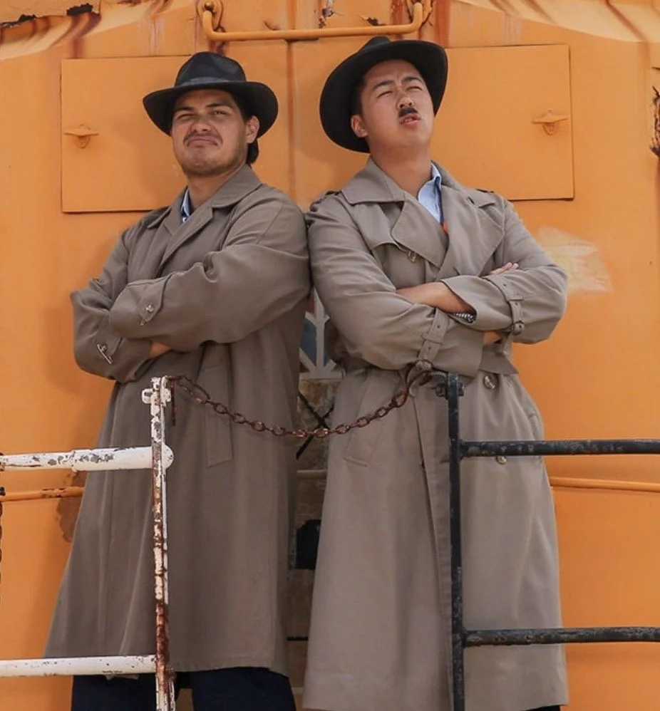 Two men dressed as explorers in trench coats and wide-brimmed hats are standing back-to-back with their arms crossed in front of an orange background.