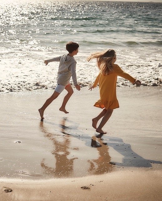 Two children, a boy and a girl, playing and running on the beach near the shoreline with ocean waves in the background.