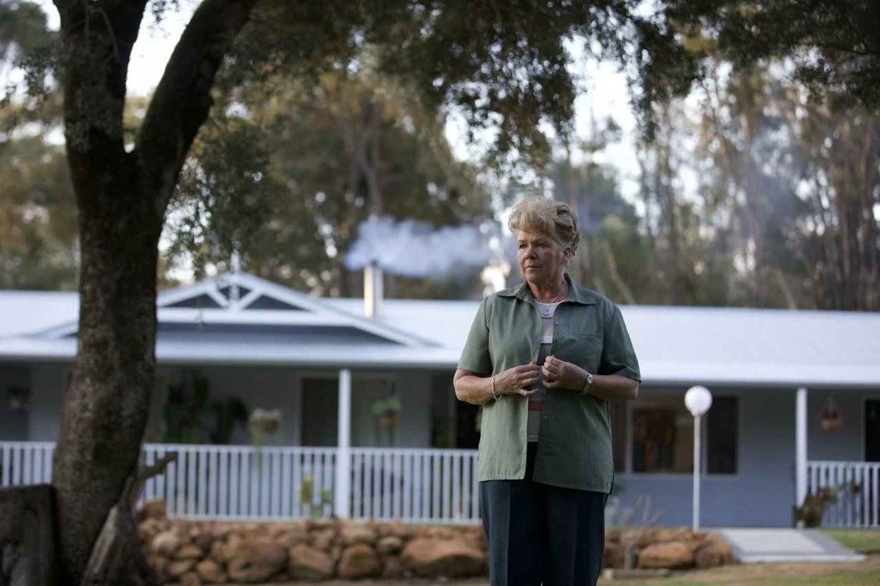 An elderly woman standing outdoors in front of a house with a porch, trees, and smoke coming from a chimney.