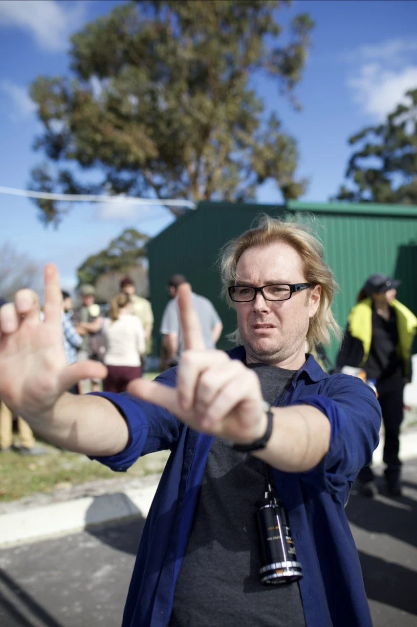 A man with blonde hair, glasses, and a blue jacket making a gesture with his hands outside on a sunny day, in front of a group of people near a green building and trees.