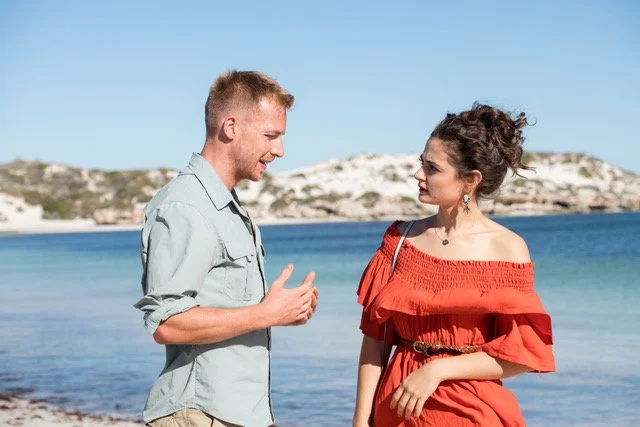A man and woman stand on a beach, engaged in conversation, with water and rocky formations in the background.