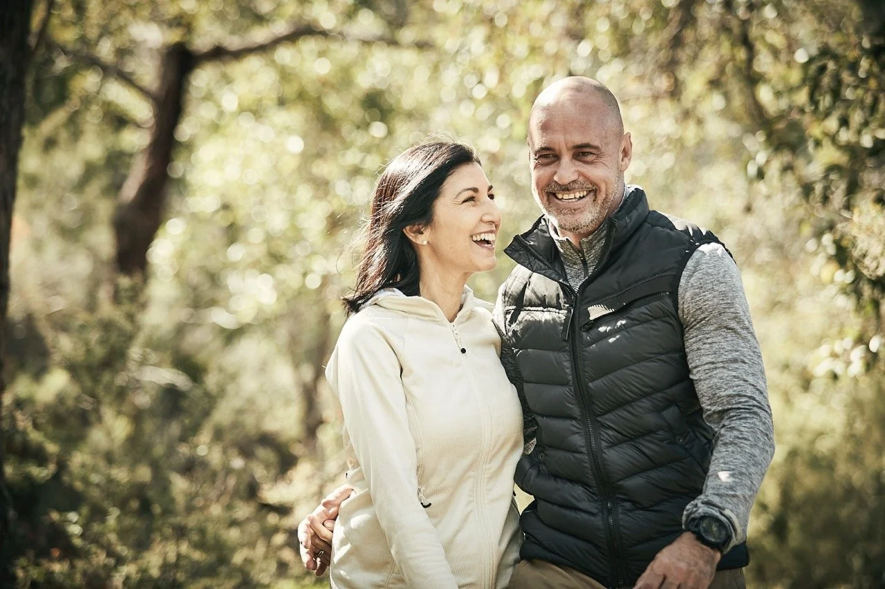 A smiling couple enjoying a walk outdoors in a forested area on a sunny day.