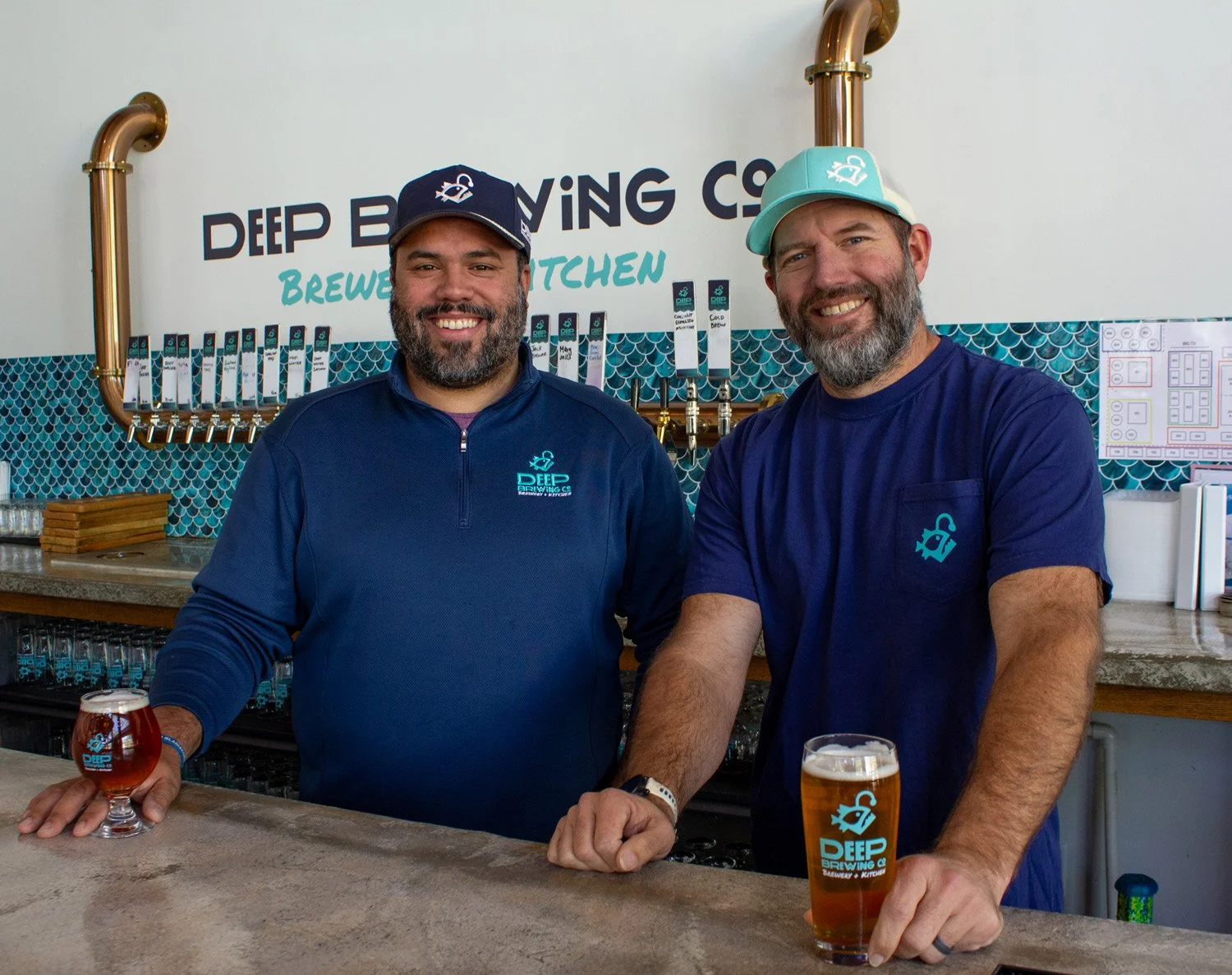 Co-Founders Justo Cruz and Ryan LaPete behind the bar counter holding glasses of beer, smiling.