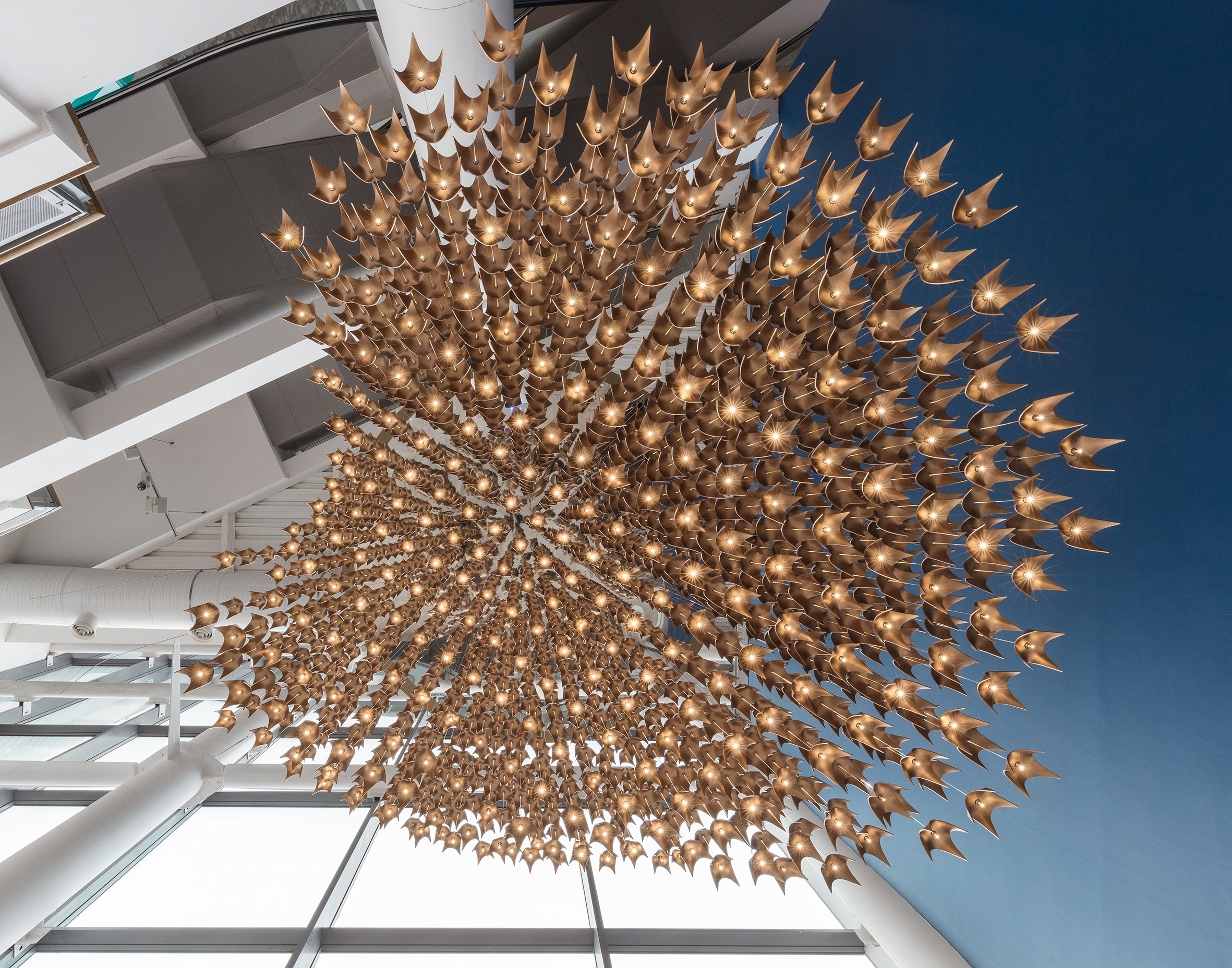 Bespoke light installation for a building lobby refurbishment in Cardiff The light installation is called Simorg (birds in flight) and is designed to reflect its surroundings and be a recognisable icon in the city. The Simorg is known as a benevolent, mythical bird in Persian mythology and often associated with the Phoenix. The Simorg was most famously described in one of our client’s favourite books, ‘The Conference of the Birds’. The cloud effect is created using parametric digital design tools. The components vary in size and were fabricated in solid cast aluminium from bespoke moulds. The light installation features 1720 brass finish components suspended via 290 fibre-optic cables. It is 6.3m high, each cable varies in length and carries 6 components. Assembling the top base of the installation on site required a bespoke scaffolding structure to be erected where 3D points were templated physically to finalise location. The base parts came pre-assembled with computer generated grid of fibre-optic cables. Mirror finish steel round plate at the top creates a further optical illusion as if the ‘birds’ continue to infinity. Design: ALEKSA studio in collaboration with Design Haus Liberty Client: Naissance Capital Real Estate Location: Cardiff Dates: 2019 Photographs by Nicholas Worley