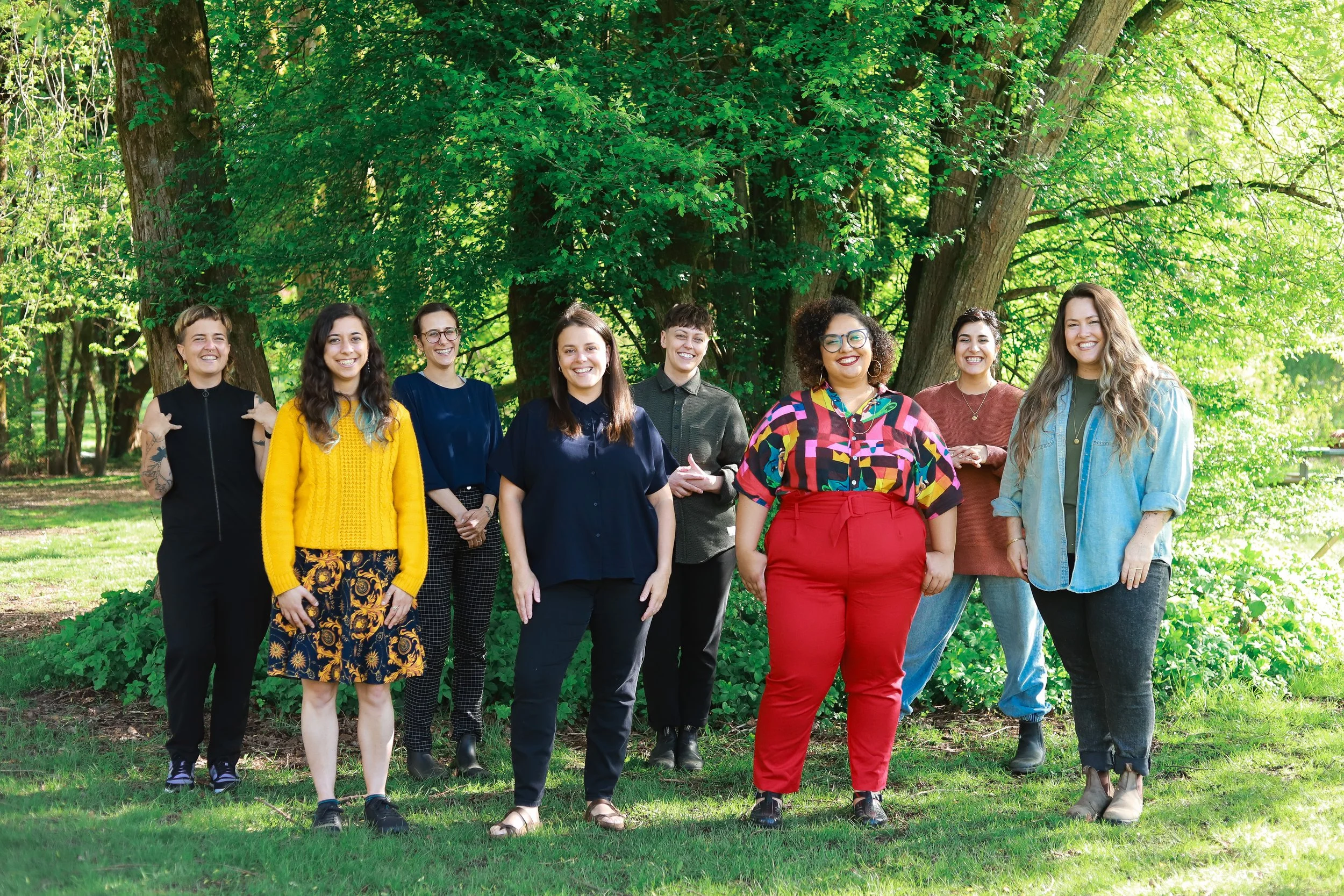 Team members stand in a group in front of trees, while looking at the camera and smiling warmly.