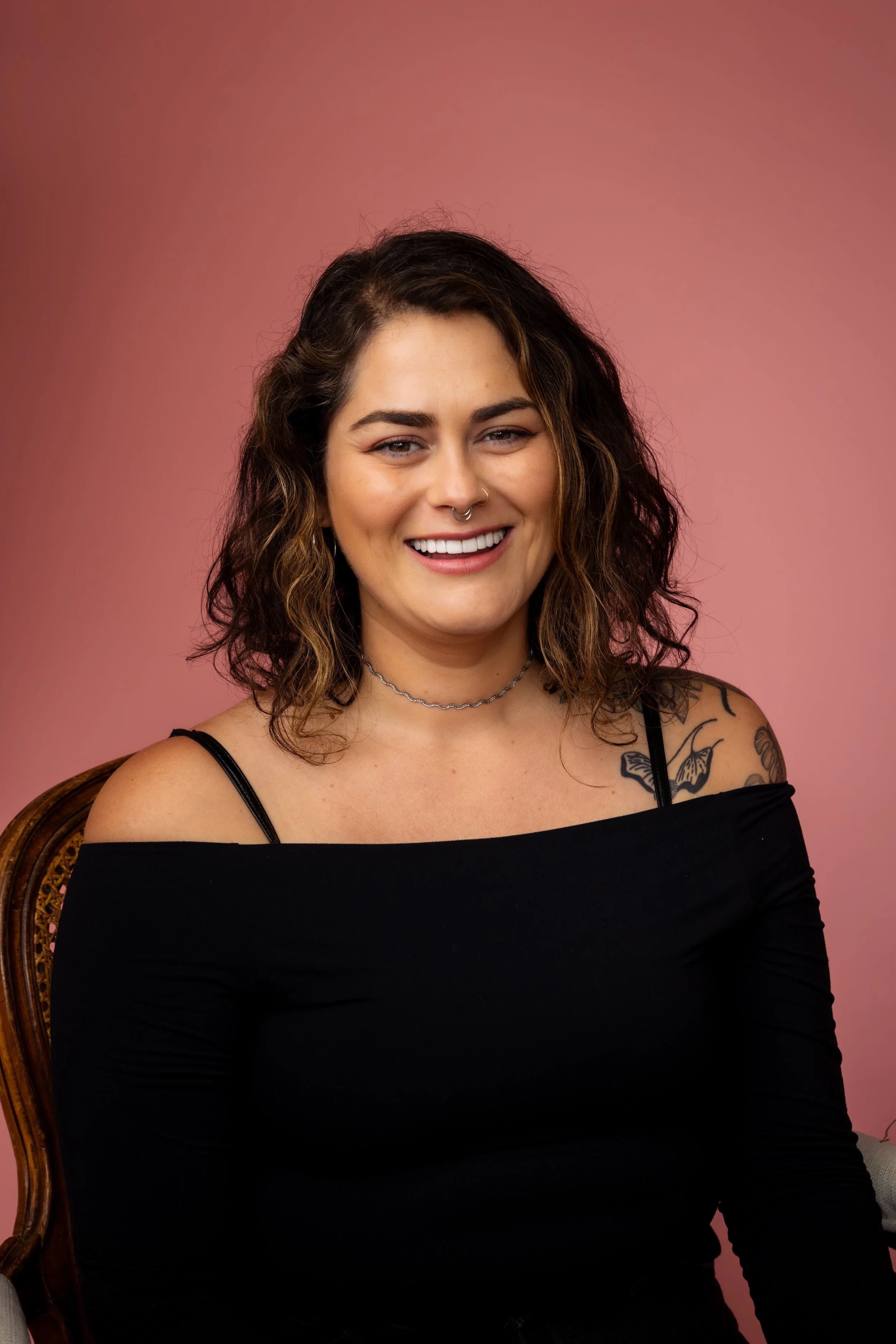 Luisa has shoulder length dark brown wavy hair and is wearing a black long sleeve top. She is seated in the middle of the frame with a coral background. She is smiling at the camera. Photo by Jade Zulkoskey.