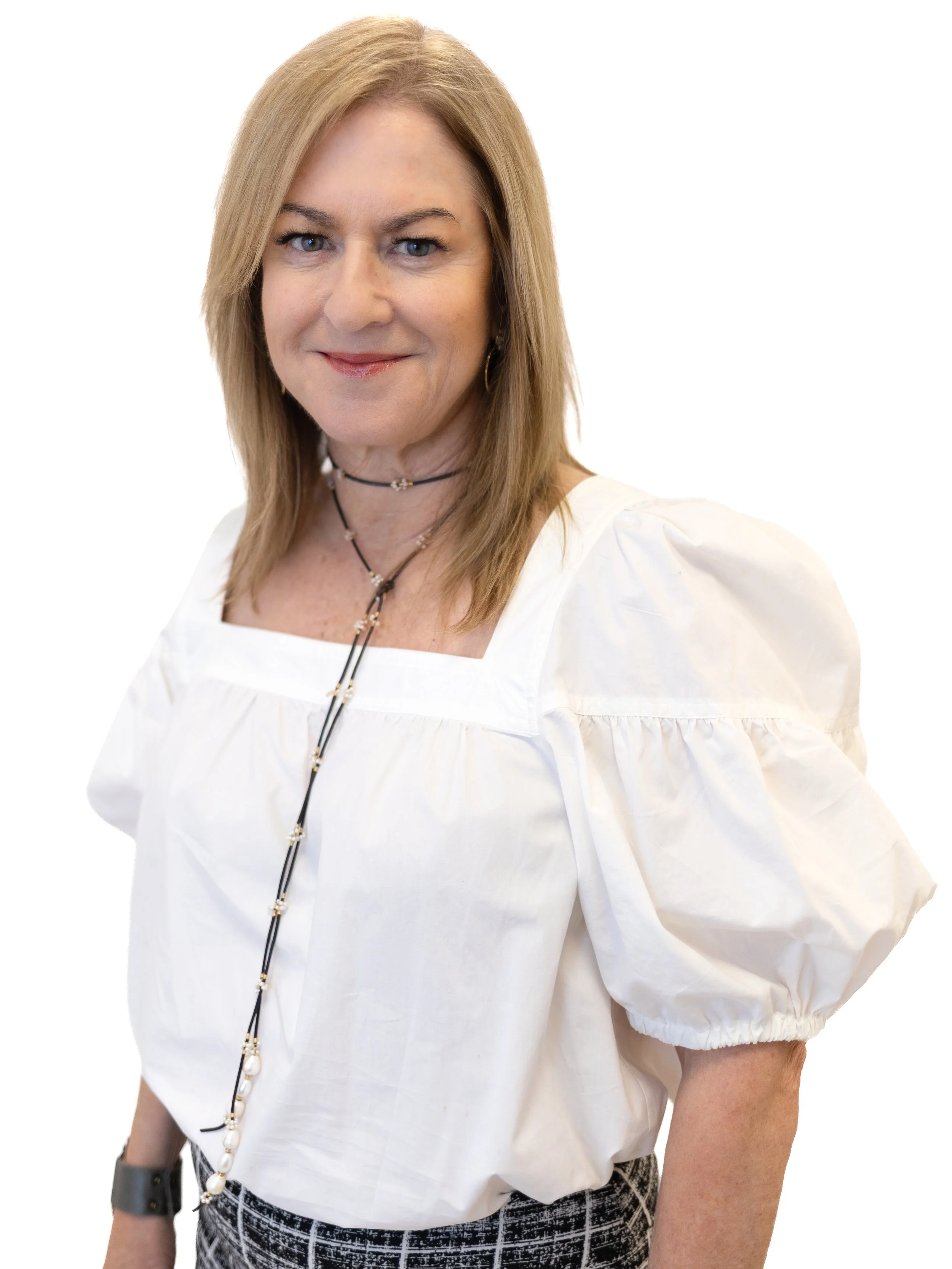 A woman with shoulder-length brown hair, wearing a white blazer, black top, and layered pearl necklaces, standing against a plain white background.