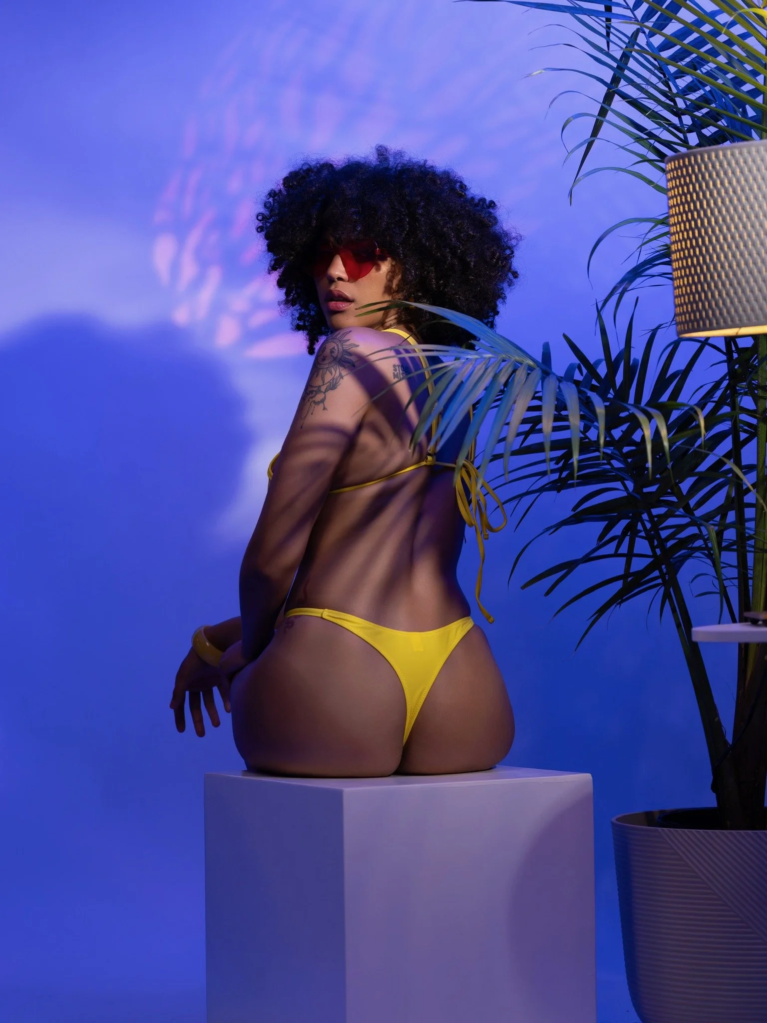 A woman with curly hair and tattoos on her arm wears red sunglasses and yellow bikini bottom, standing atop a white pedestal in front of a blue background, surrounded by green tropical plants and a tall textured lamp.