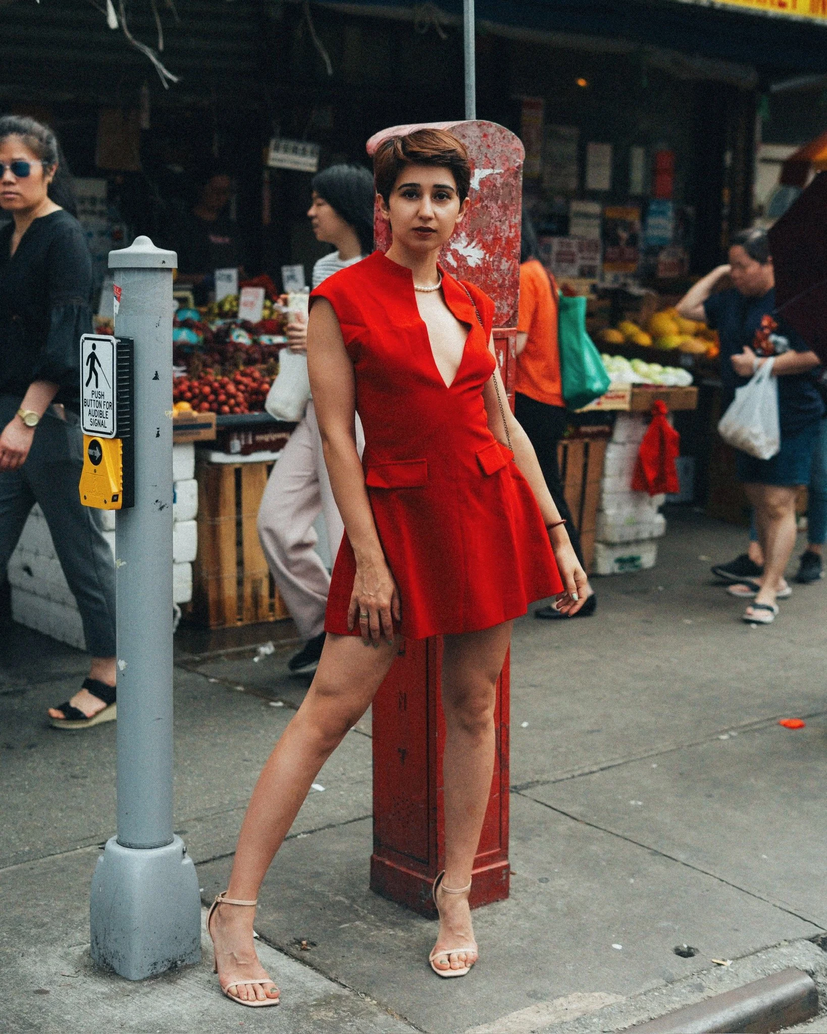 A woman wearing a red dress and beige high-heeled sandals standing on a city sidewalk next to a red fire hydrant and a parking meter, with other pedestrians in the background.