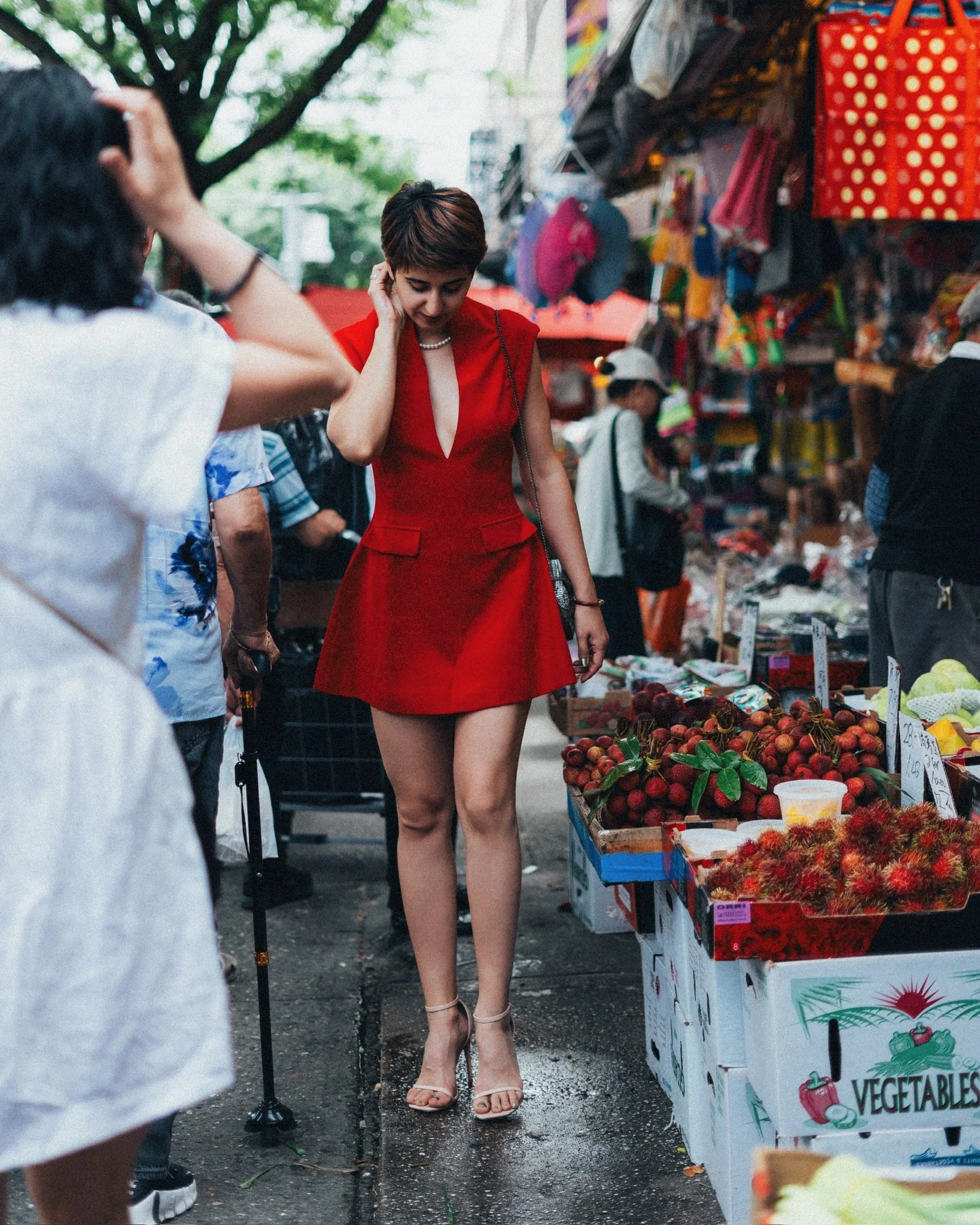 A woman with short hair in a red dress shopping at an outdoor vegetable market, surrounded by colorful produce and other shoppers.