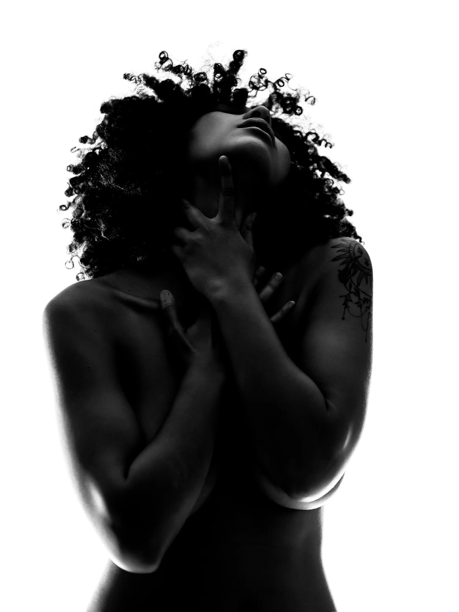 Black and white photograph of a woman with curly hair touching her chin and chest, with a tattoo on her shoulder, against a plain white background.
