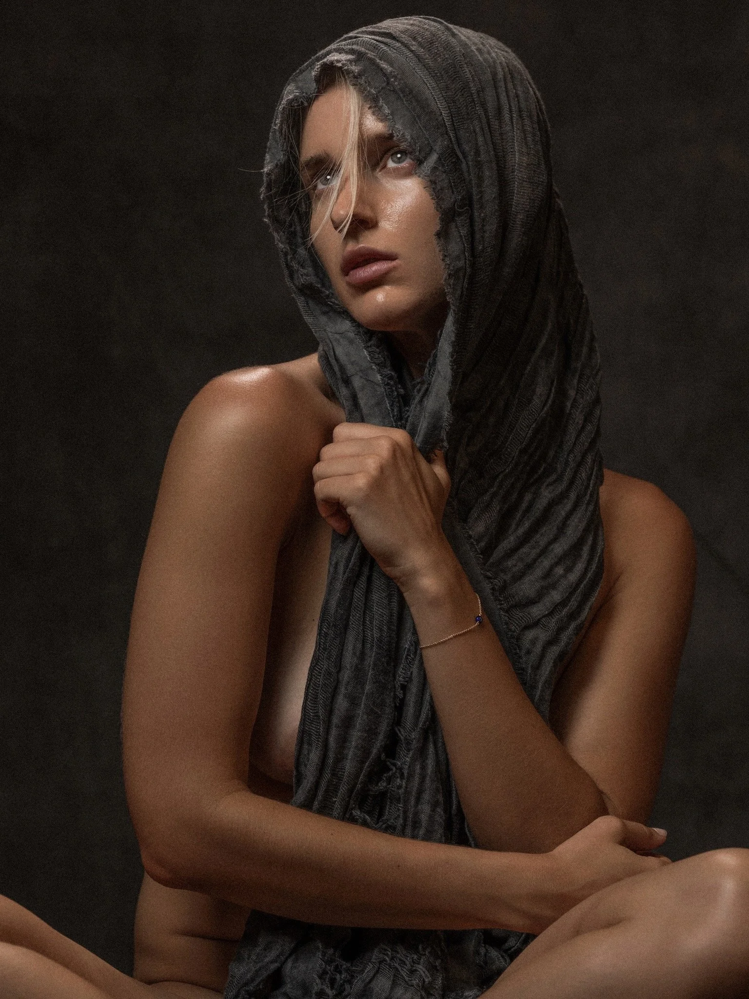 A woman with light skin is wrapped in a black, textured hooded scarf, holding it with her right hand, against a dark background. She has blonde hair peeking out from under the hood, and her gaze is directed upward with a subtle, thoughtful expression